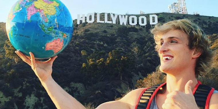 Logan Paul holds his hand in a shaka while holding a globe in front of the Hollywood sign.