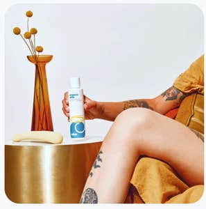 A tattooed woman holds Cake's Organic Aloe lube in her hand with a vibrator and plant on the end table next to her.