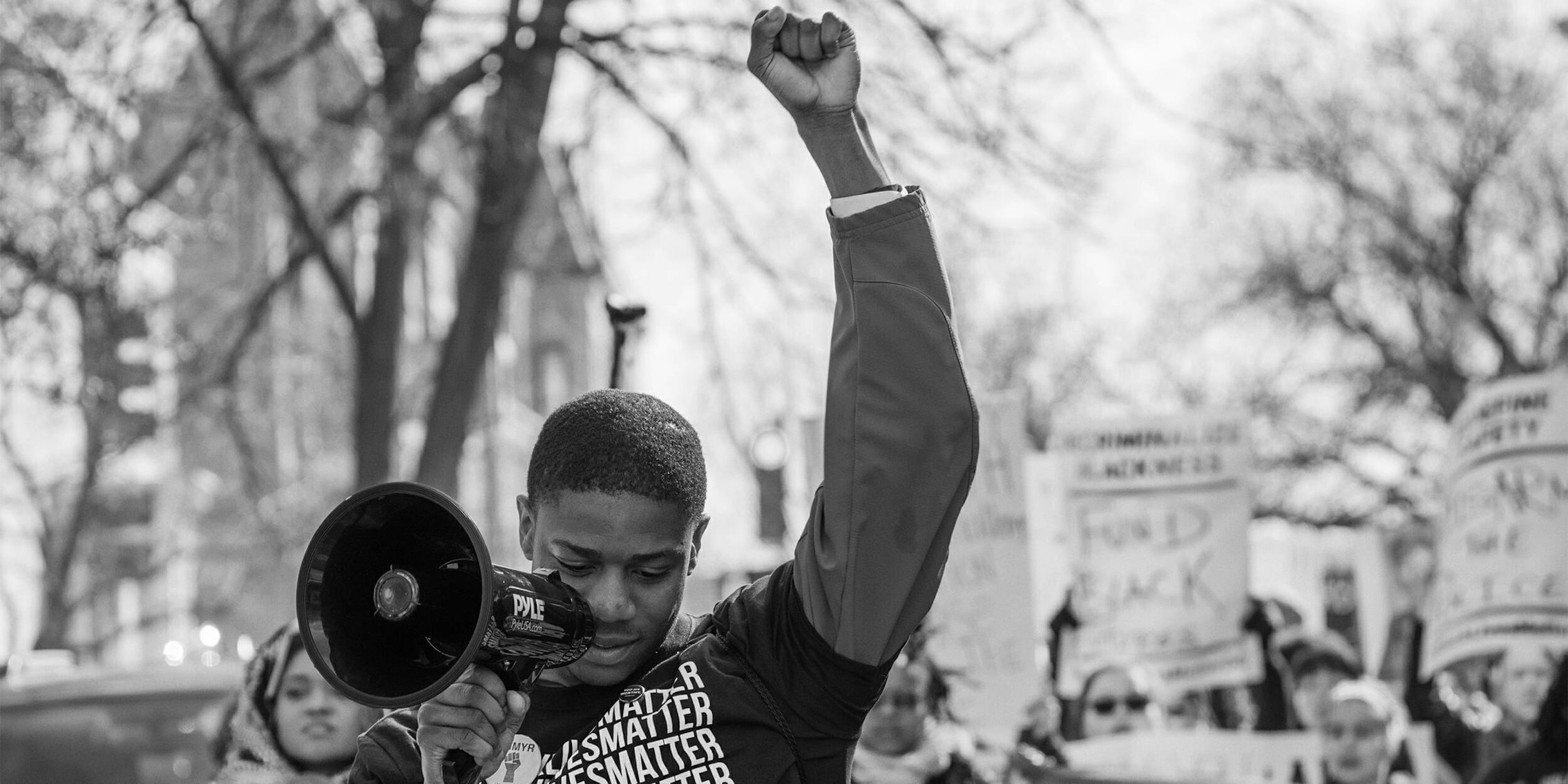 Man in 'Black Lives Matter' shirt raising fist, speaking through megaphone