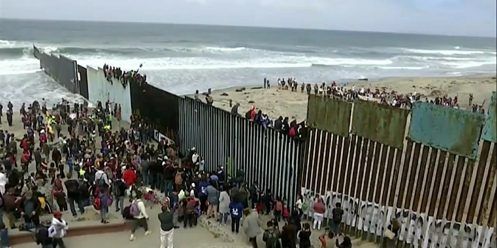 A caravan of Central American migrants standing along and sitting on the U.S.-Mexico border wall in San Diego.