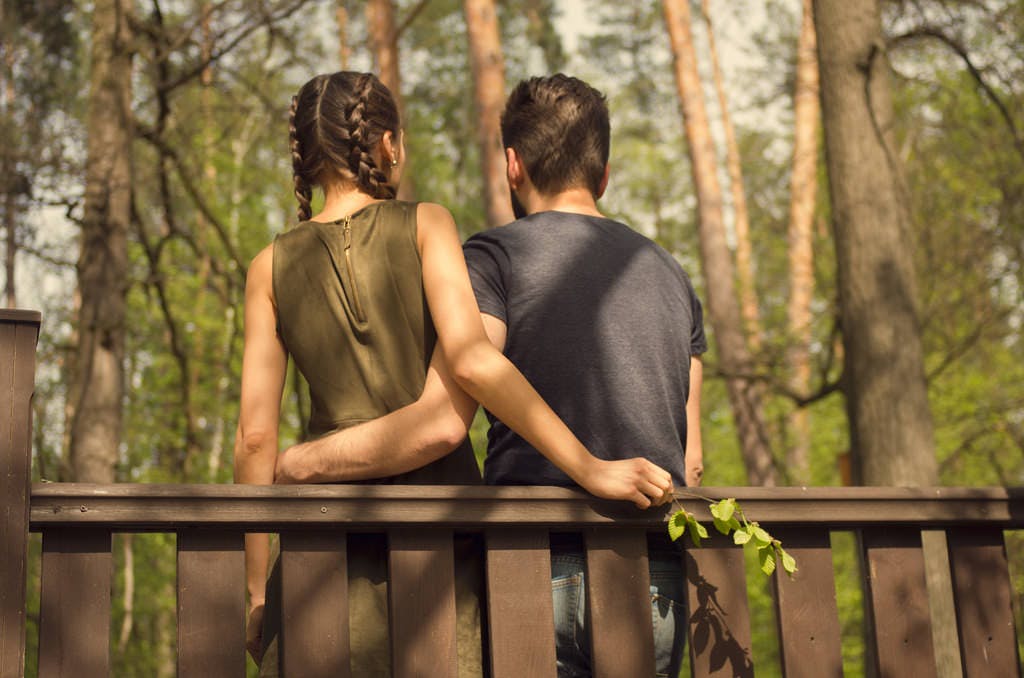 How to DTR - a photo of two people leaning casually up against a deck railing, their backs facing the camera, their arms lightly embracing each other around their waists, both facing a wooded area. The person on the left is in a green sleeveless dress and wearing braids, the person on the right has short-cropped hair and is wearing a dark navy t-shirt and jeans.
