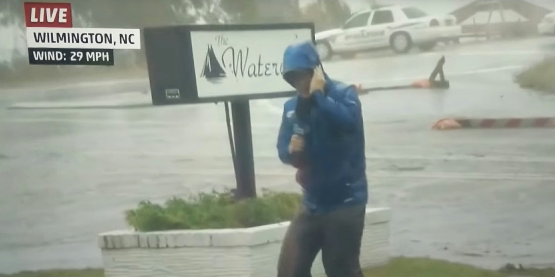 Weatherman dramatically braces against Hurricane Florence winds as dudes stroll casually in the background.