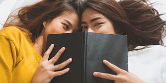 two women laying in bed, smiling behind book