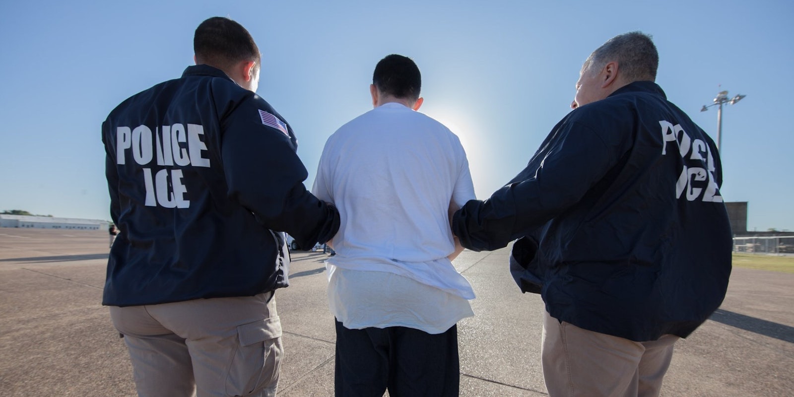 Two ICE officers are seen from the back arresting a man in a white t-shirt
