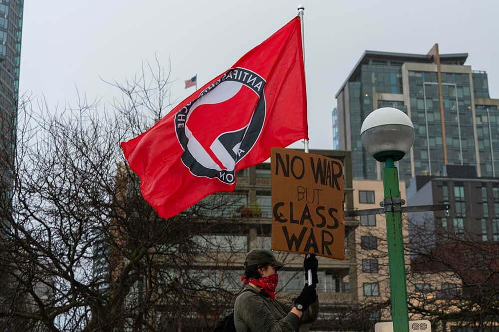 protester holds antifacist flag and "no war but class war" sign