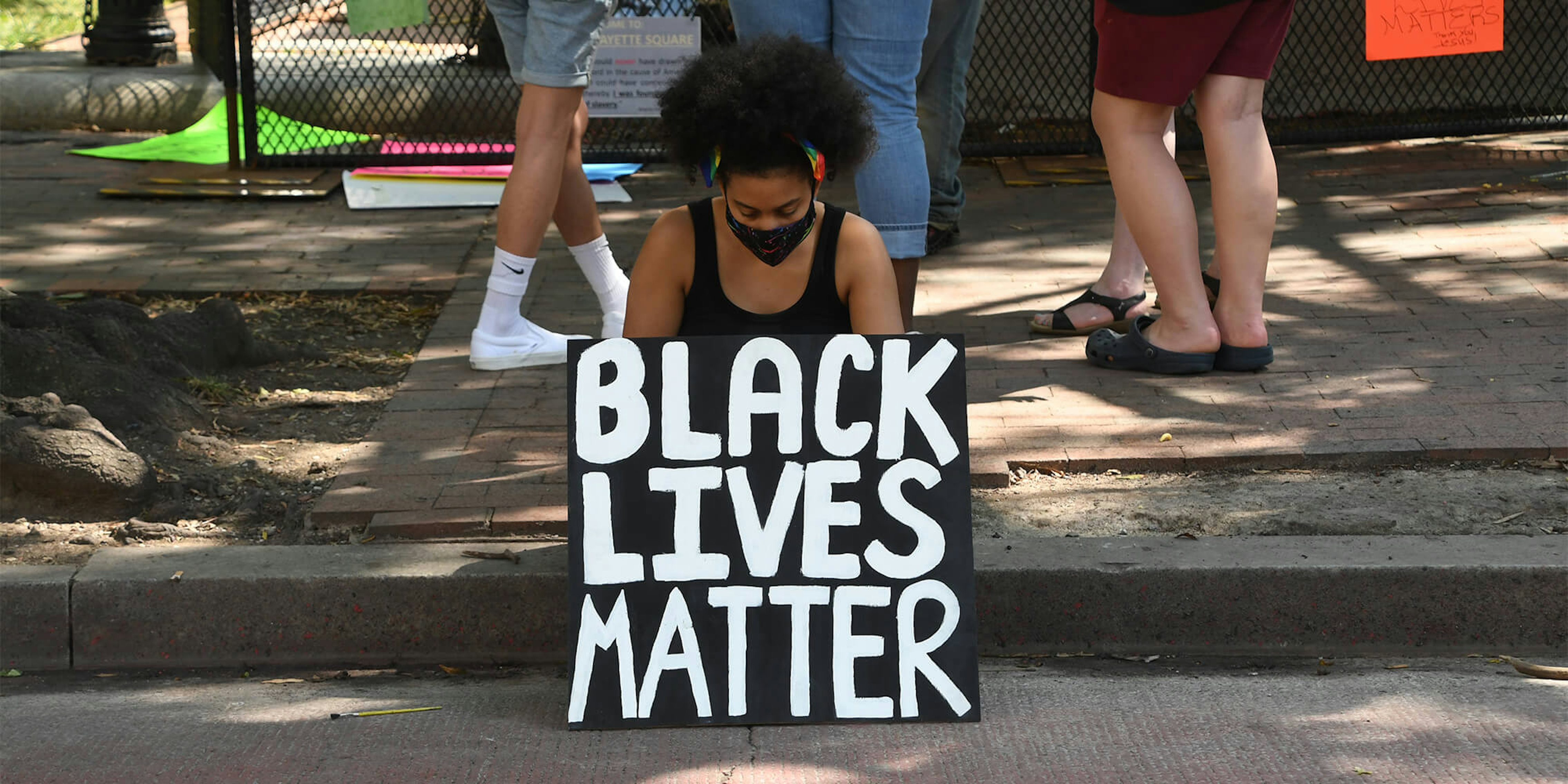 woman sitting on curb holding a Black Lives Matter sign, wearing a facemask