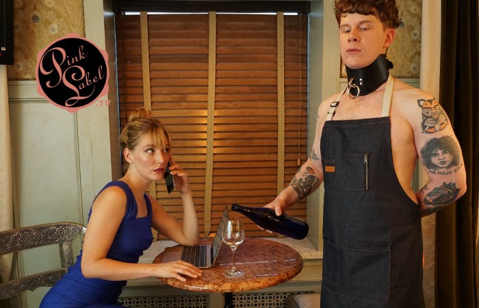 An image showing a woman working on her laptop and phone, while a tattooed man in an apron and large collar pours her a glass of wine.
