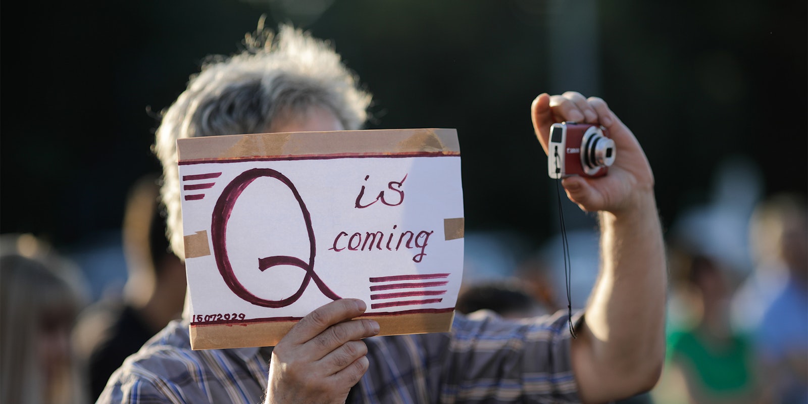 man holding camera and 'q is coming' sign