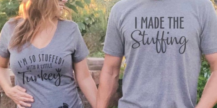 a white pregnant woman with wavy blonde hair wearing a grey t-shirt saying 'I'm so stuffed with a little turkey' and a pair of baby footprints, she's smiling at a man in a matching shirt that says 'I made the stuffing'