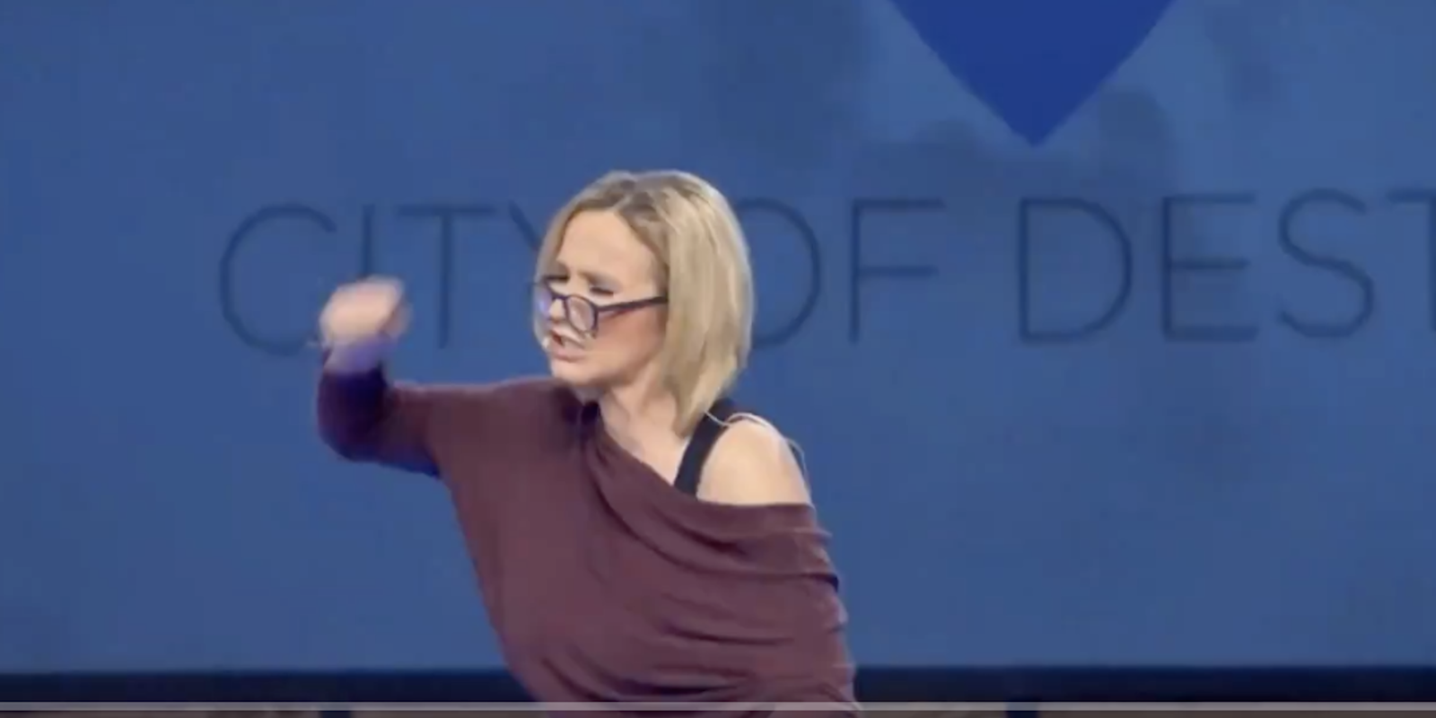Paula White-Cain with her face turned to the left and her fist raised in the air. She has a blonde bob and is wearing a purple off the shoulder top.