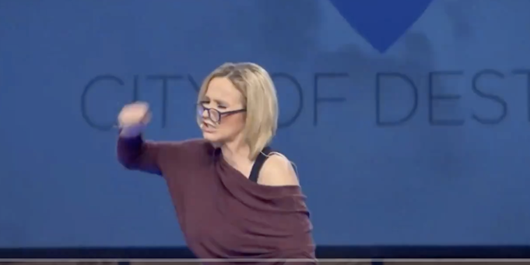 Paula White-Cain with her face turned to the left and her fist raised in the air. She has a blonde bob and is wearing a purple off the shoulder top.