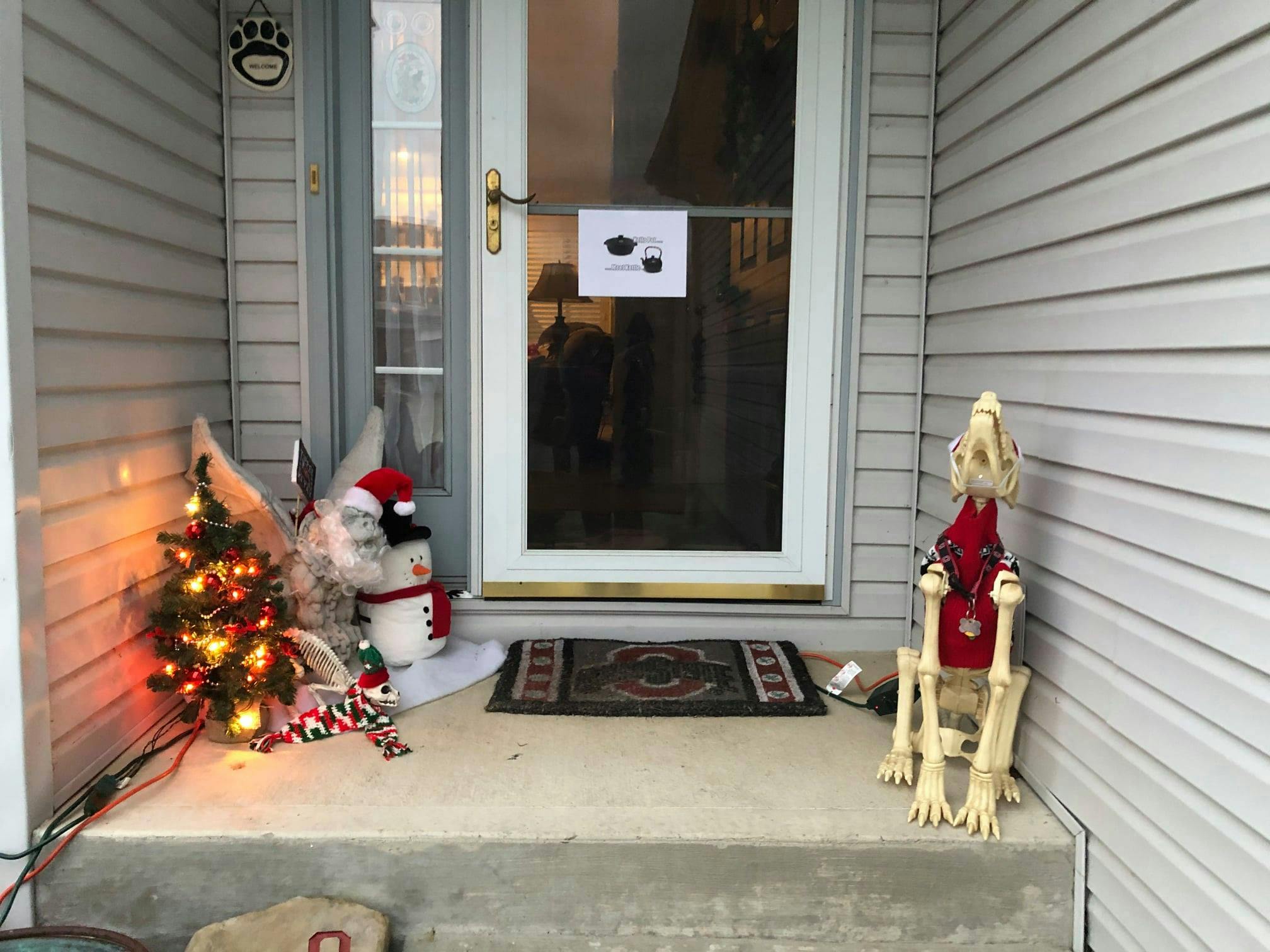 Frank the Gargoyle, the small Christmas tree now covered in multicoloured lights, the elf sitting on Frank's shoulder, a tilted placard where the image of Bruce Willis is hard to see and a plastic cat skeleton in a Christmas sweater