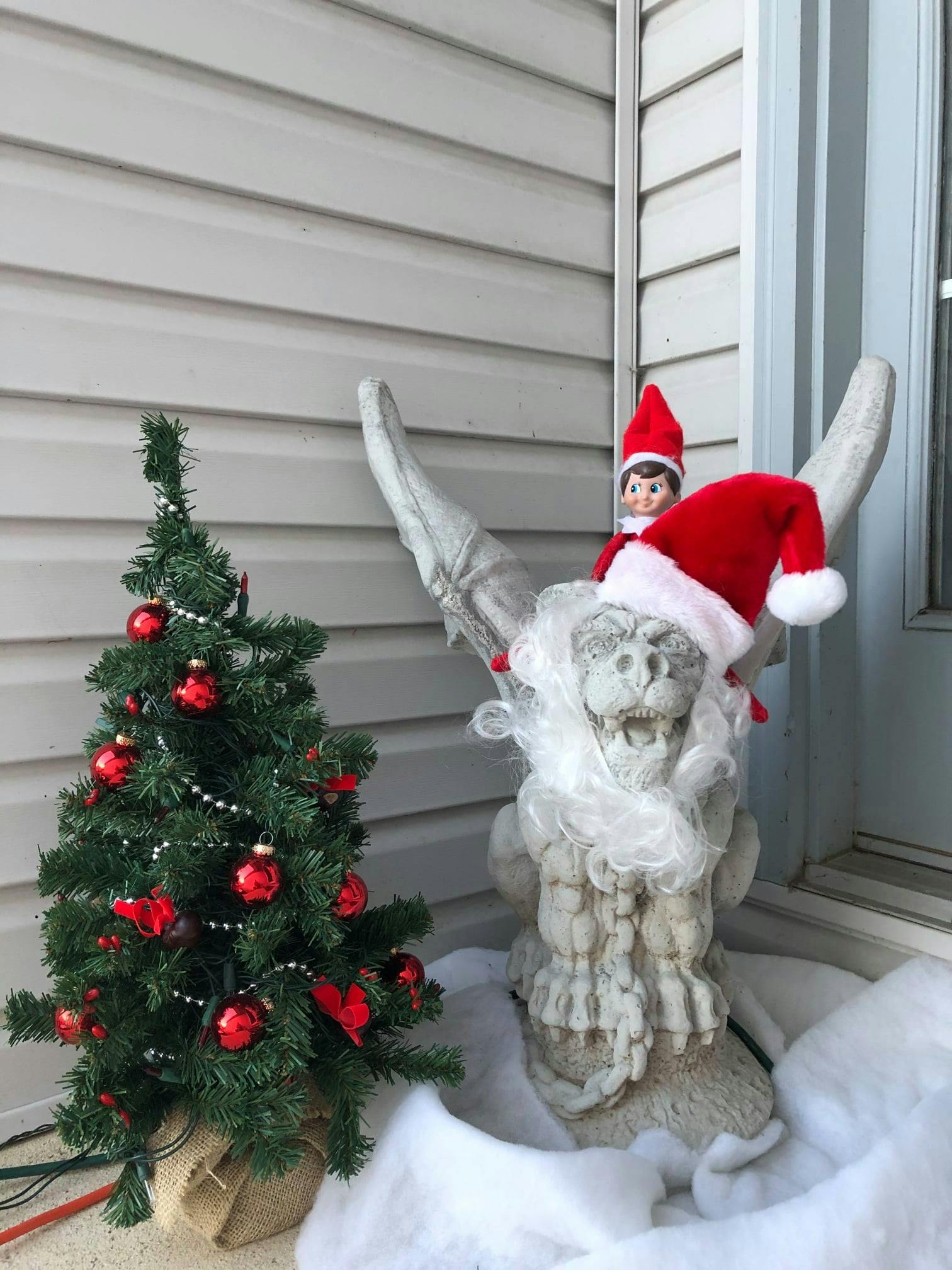 The gargoyle wearing a Santa hat and beard next to a small Christmas tree with red baubles. An elf on the shelf is sitting on the gargoyle's shoulder