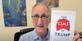 an older white man in black framed glasses makes an awkward, earnest expression while holding up a red and white book called The Cult of Trump