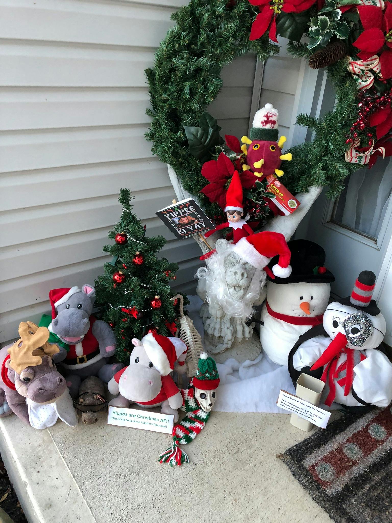 three hippos in Santa clothing, a small Christmas tree with red ornaments, Frank the Gargoyle, two plushie snowmen, an elf on the shelf holding a Die Hard placard and a Christmas hat wearing coronavirus plushie coming out of a wreath