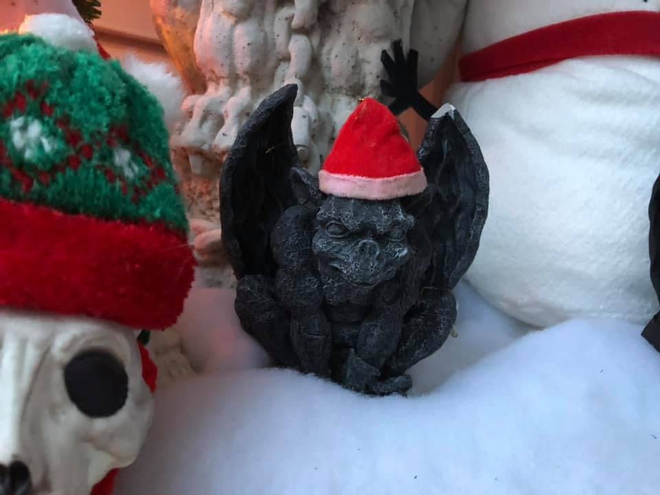 A little black stone gargoyle wearing a Santa hat crouches in the fake snow, part of the skeletal cats skull and the hat it's wearing can be seen on the left