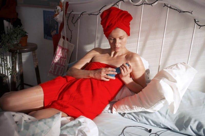Photo of a woman laying in bed holding a rabbit vibrator with a red towel in her hair and another wrapped around her body.