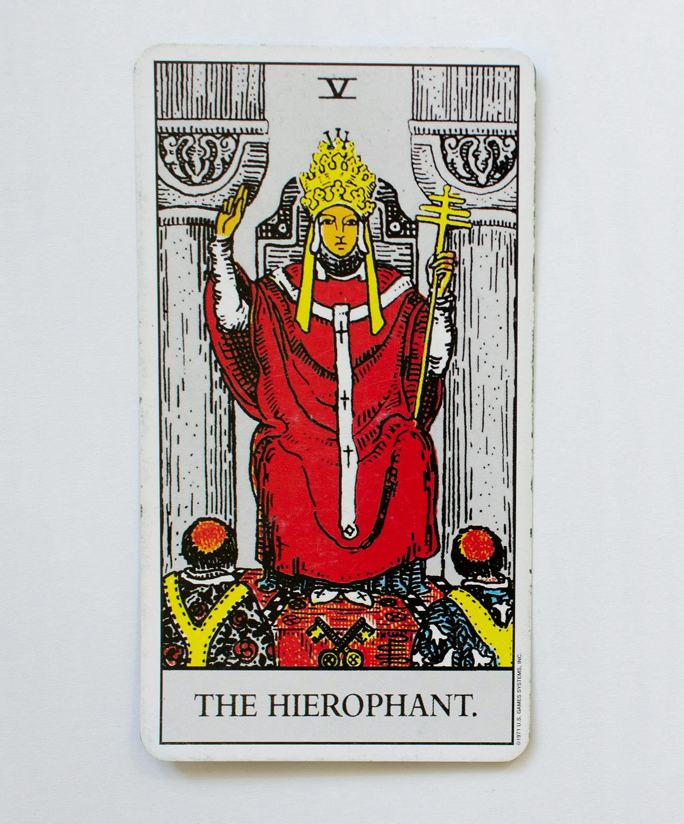 The Hierophant tarot card. Image of a person sitting in a throne between two pillars wearing a crown and red cape, with both hands raised one holding a scepter.