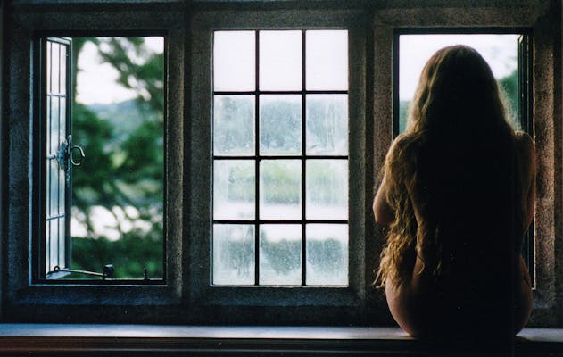 A naked woman with long hair sits silhouetted in front of a window.