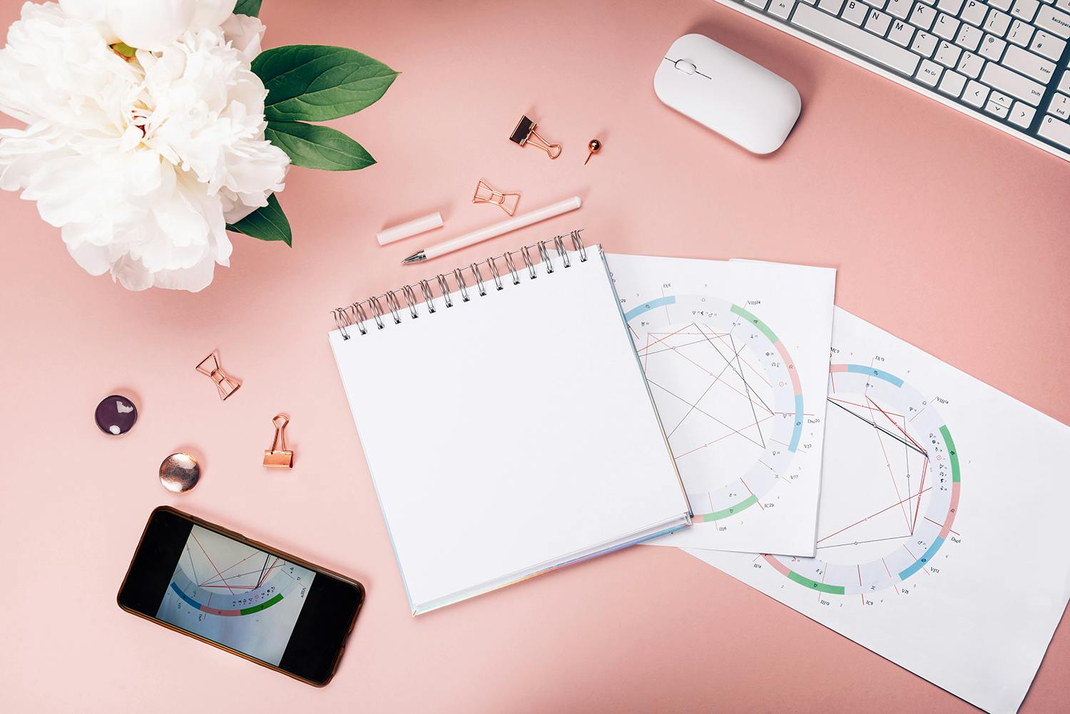 Photo of a cellphone, notebook, hand drawn birth charts, and a computer with a bouquet of flowers next to it on a pink background.