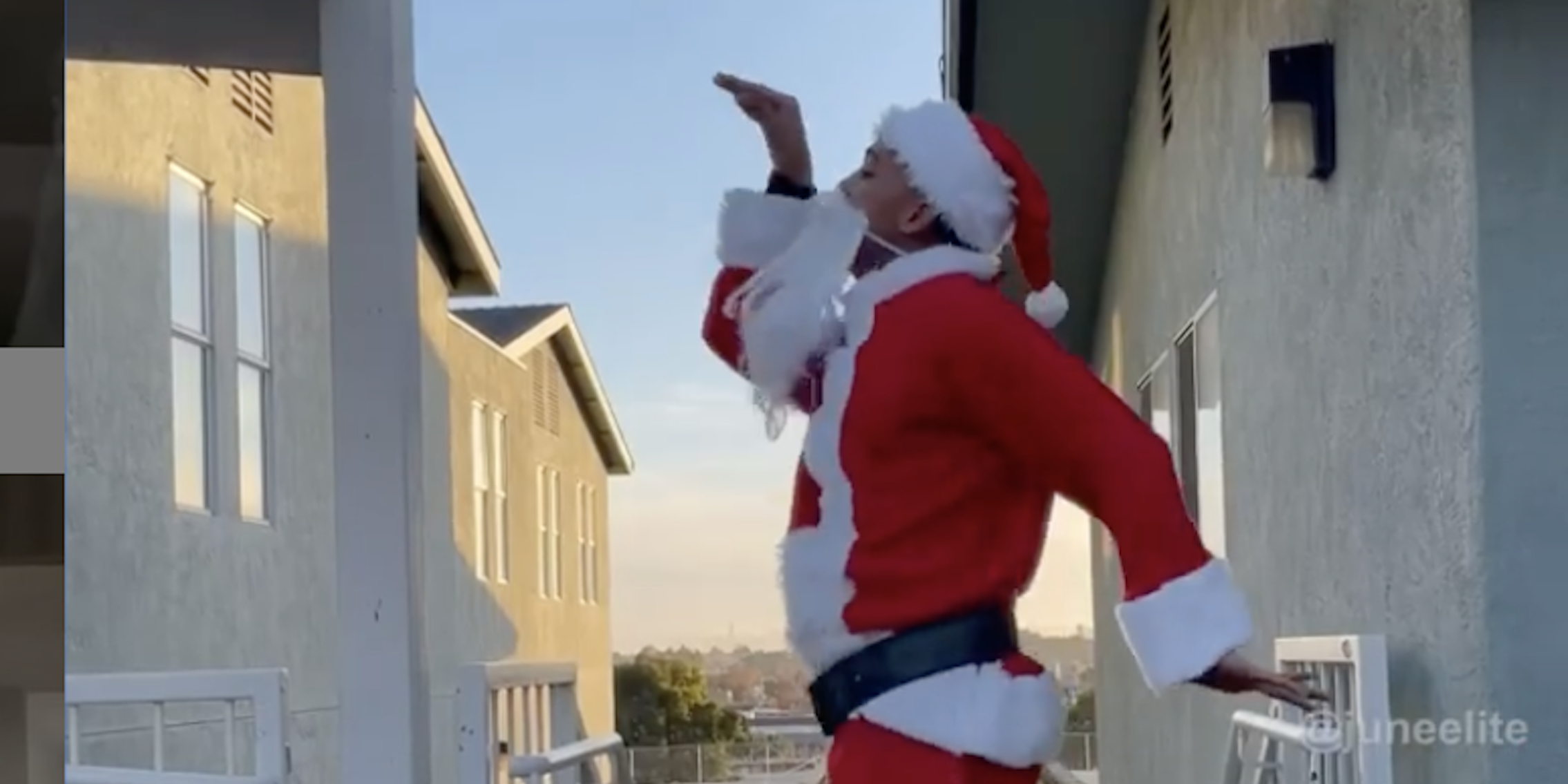A Black man in a Santa suit and beard does the Junebug Challenge dance in an alley between houses