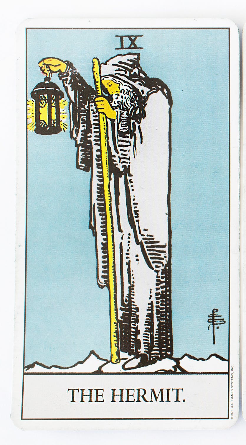 The Hermit in the Ride-Waite deck. Features an image of a man dressed in white holding a walking stick in one hand and a latern in the other.