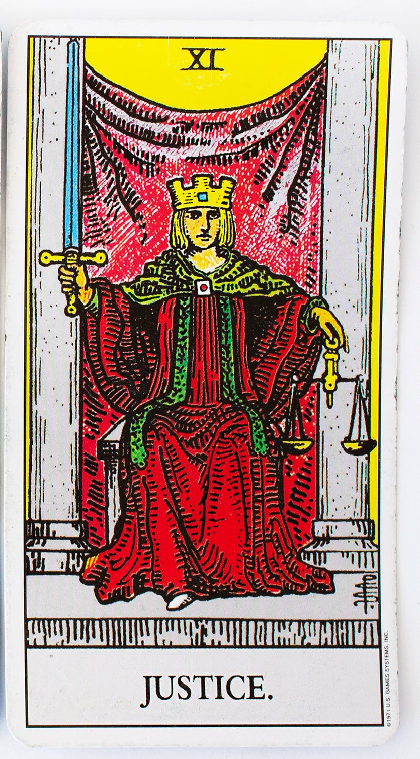 The justice card from the rider-waite deck. Features an image of a person sitting in a throne wearing a red robe and a crown, holding a set of scales in one hand and a sword in the other.