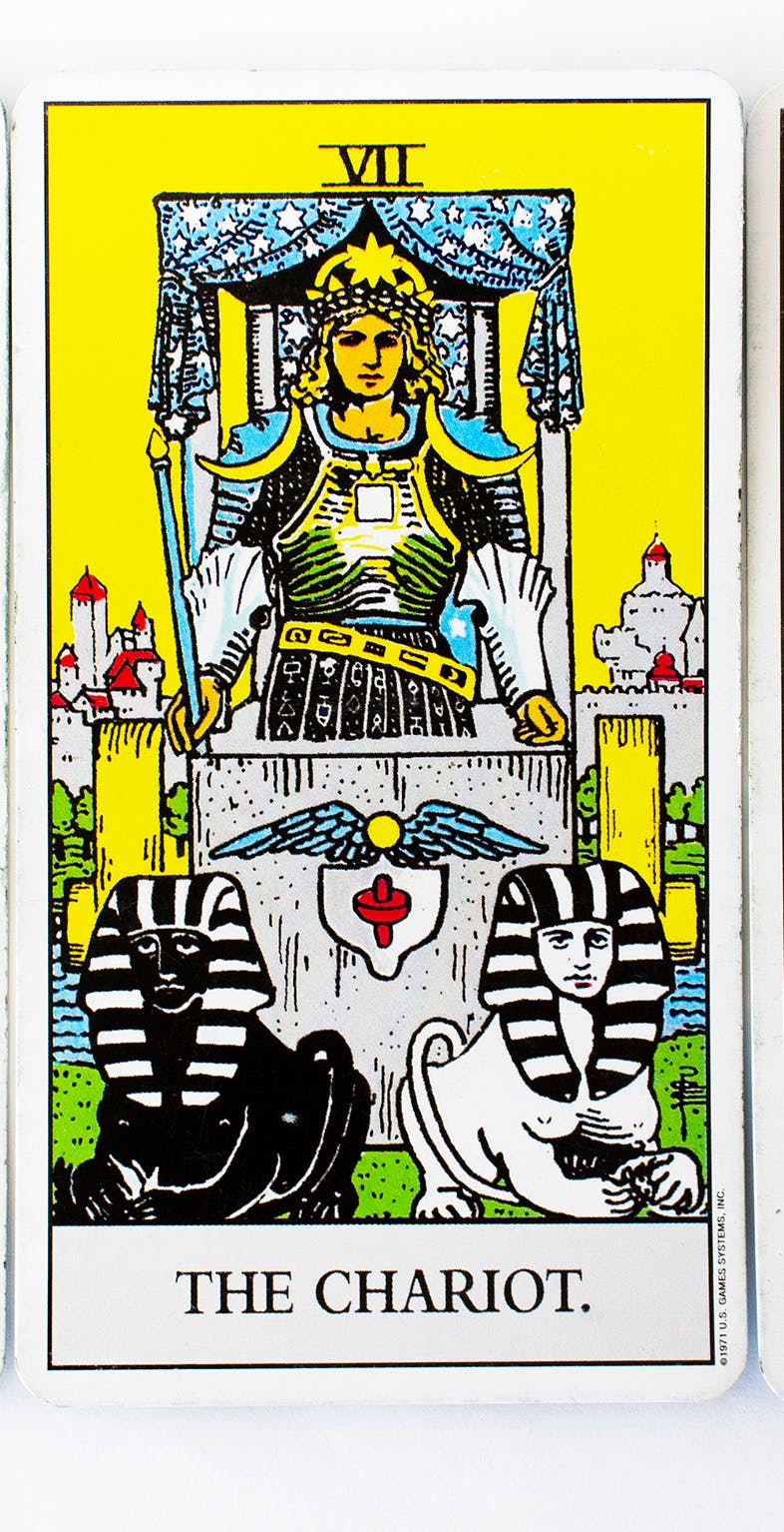 The Chariot card in the Rider-waite tarot deck. Image of shows a brave warrior standing inside a chariot. He wears armour decorated with crescent moons and has two lions at his feet.