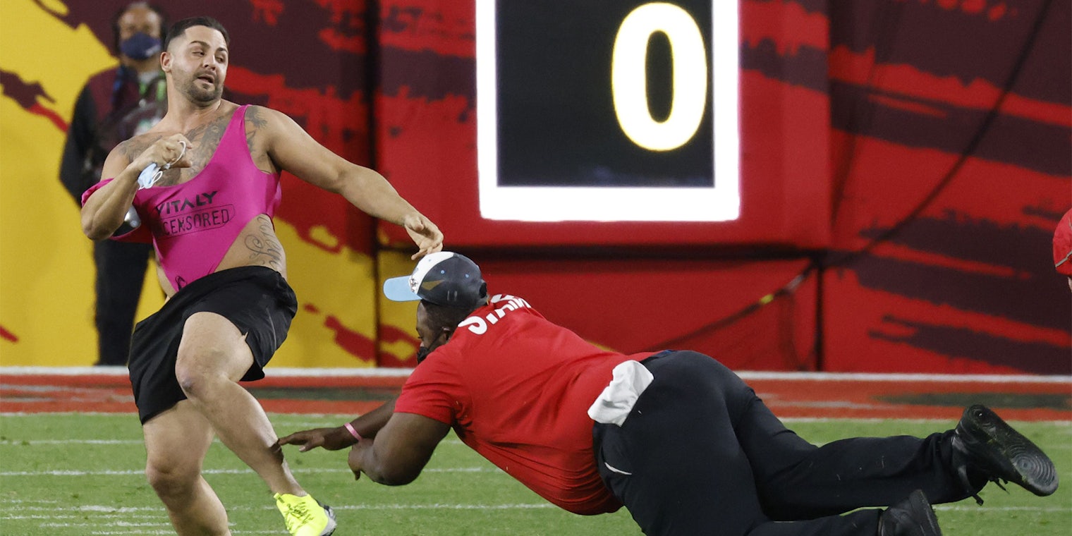 Security guard dives toward fan in pink onesie and black shorts running across the field of Super Bowl LV.