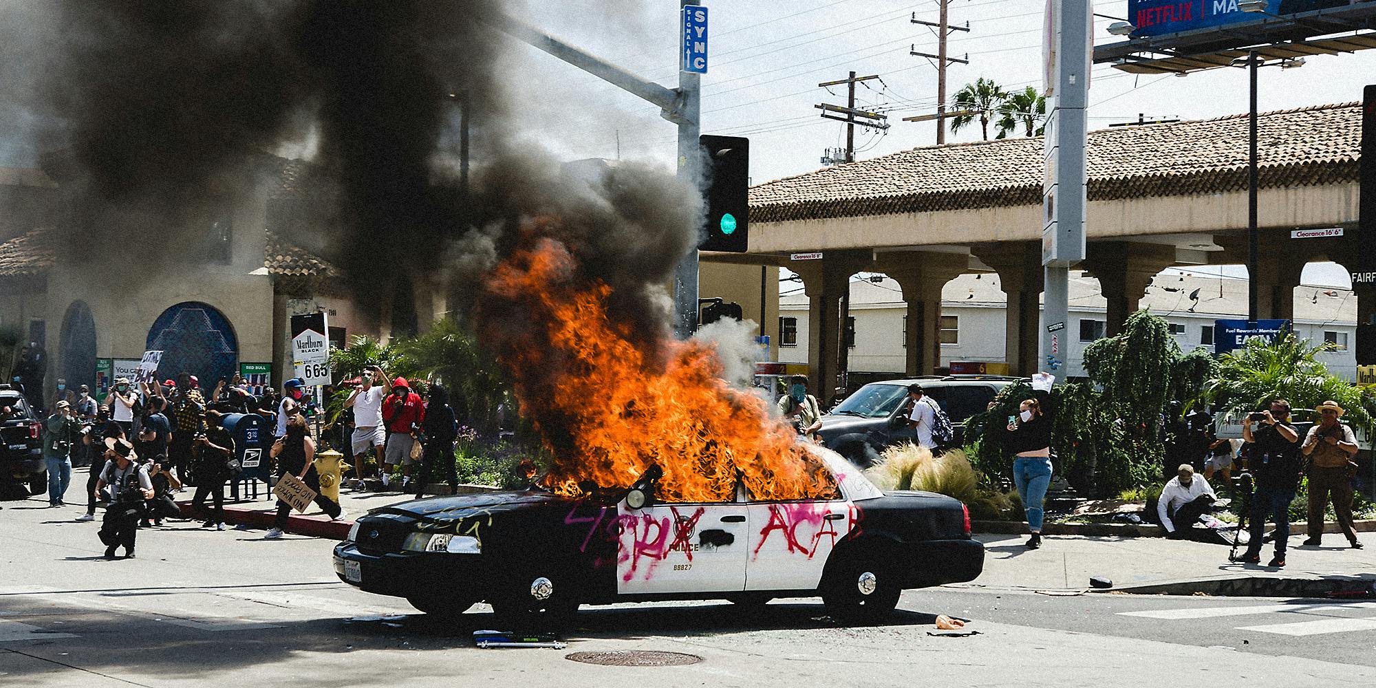 An LAPD car on fire while people watch it burn.