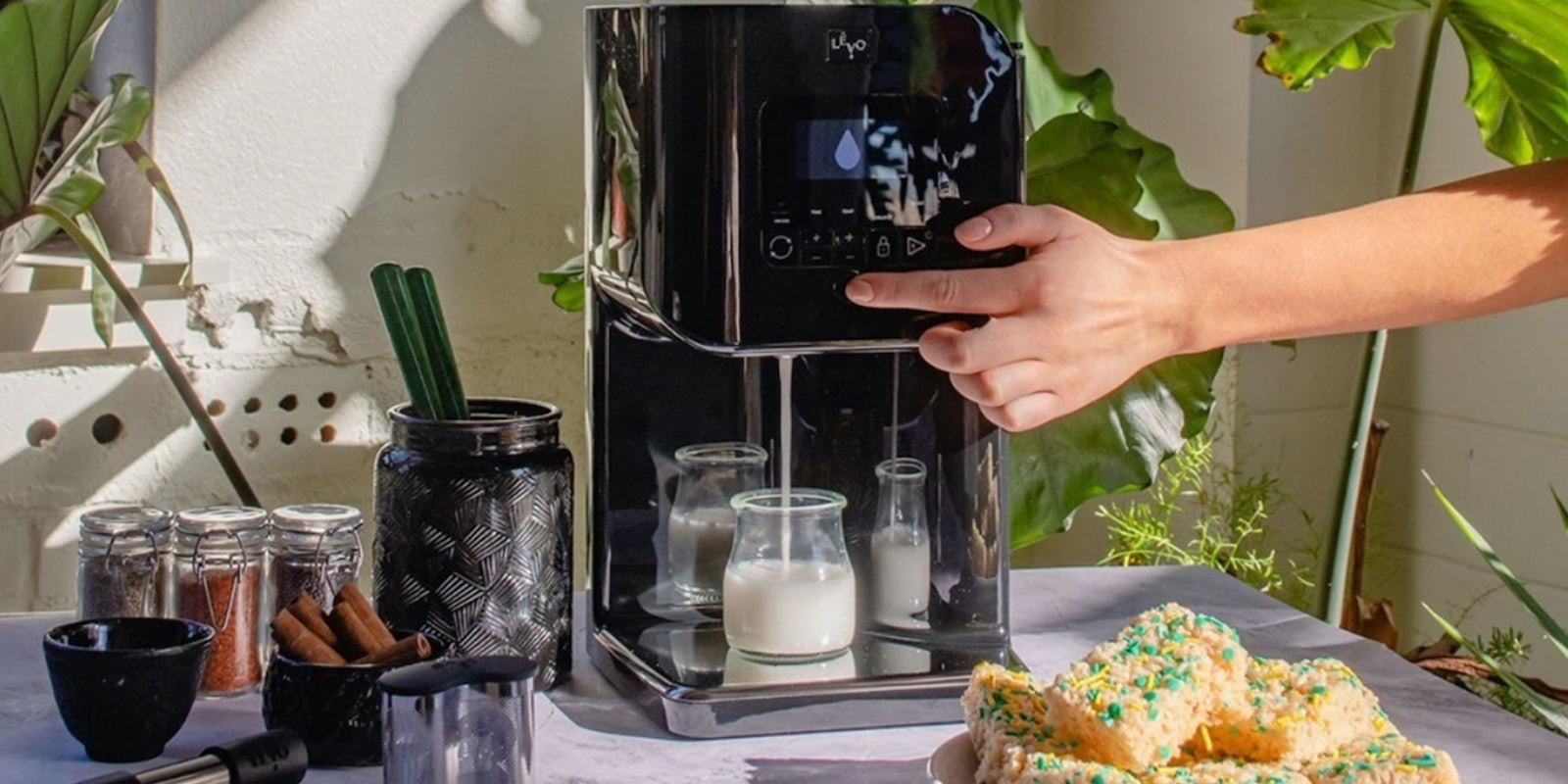 LĒVO II cannabutter machine is busy dispensing infused milk on a table next to the device's accessories, rice krispy treats, and a plant in the background.