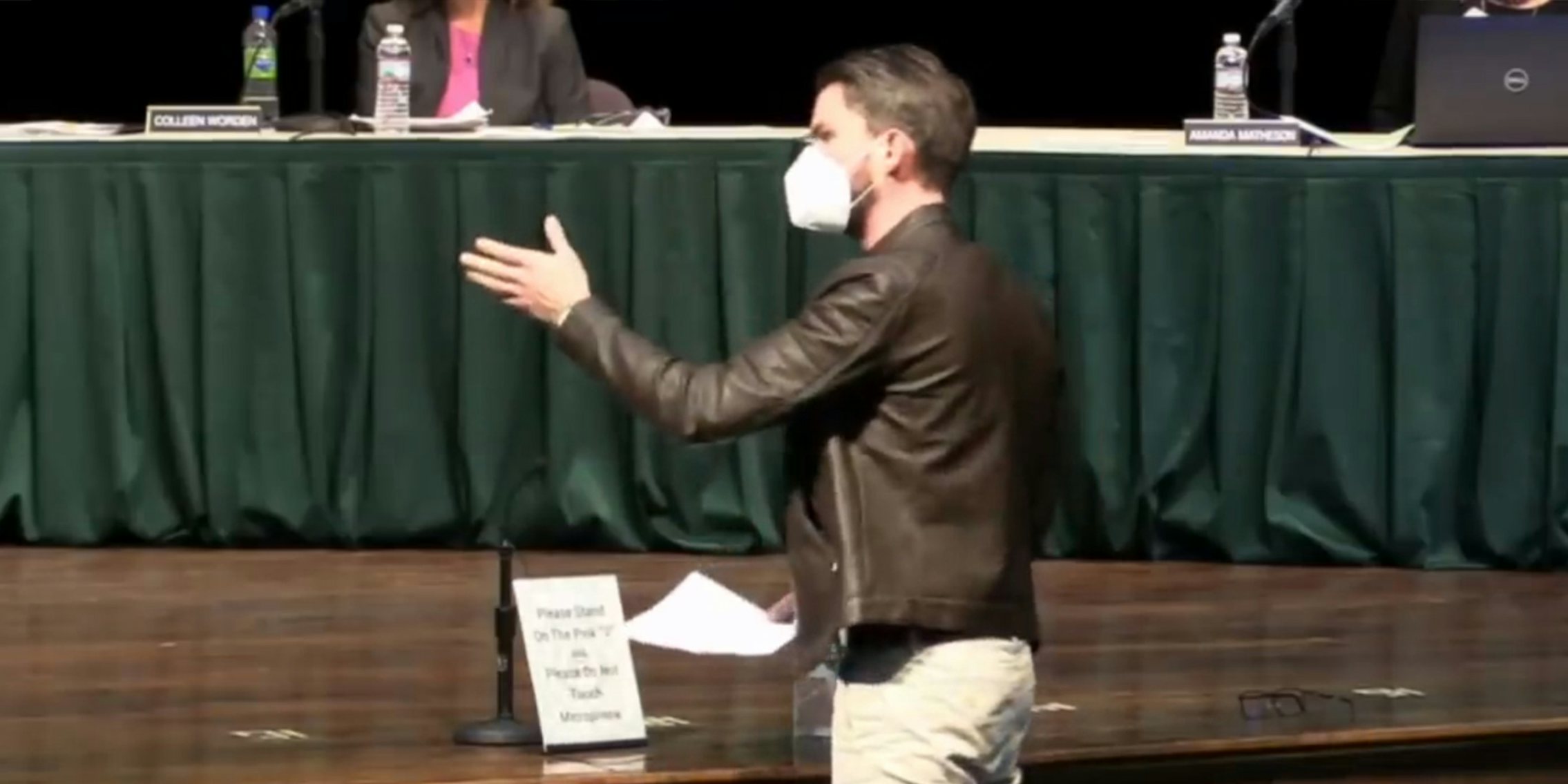 man in leather jacket and mask gestures to people sitting at table on stage