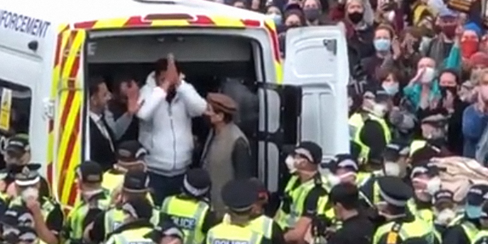 man holding hands together in thanks while exiting the back of a police van, surrounded by police and neighbors