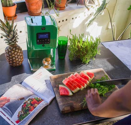 LĒVO II on a counter next to a pineapple, a cookbook, some greens as someone chops watermelon and herbs on a cutting board.