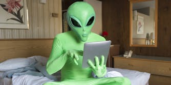 Green alien sitting using tablet computer on the bed in a hotel room