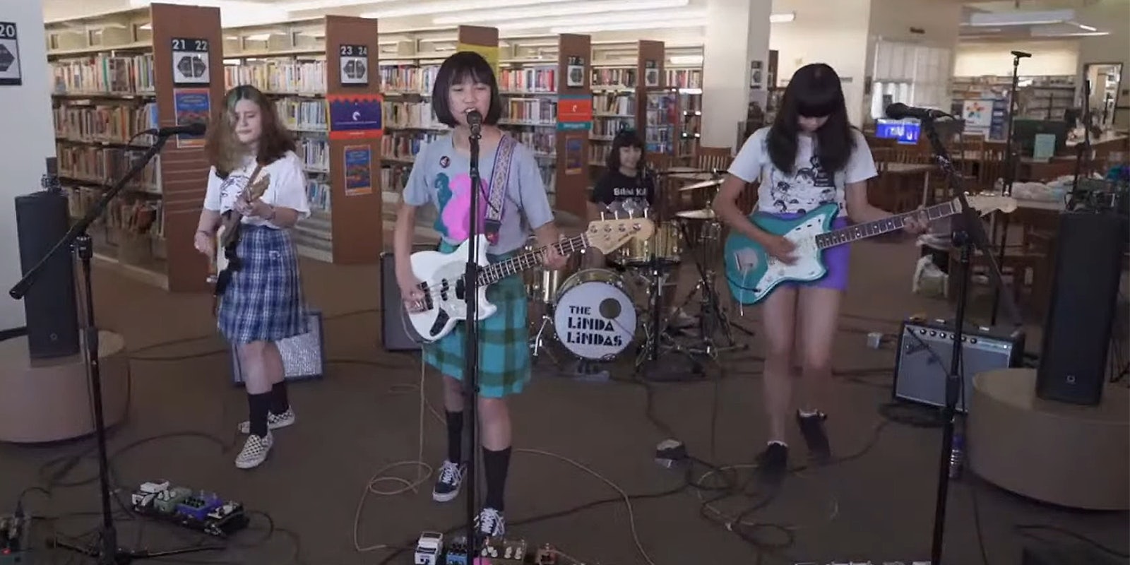 Four young girls perform on bass, guitars, and drums in a public library.