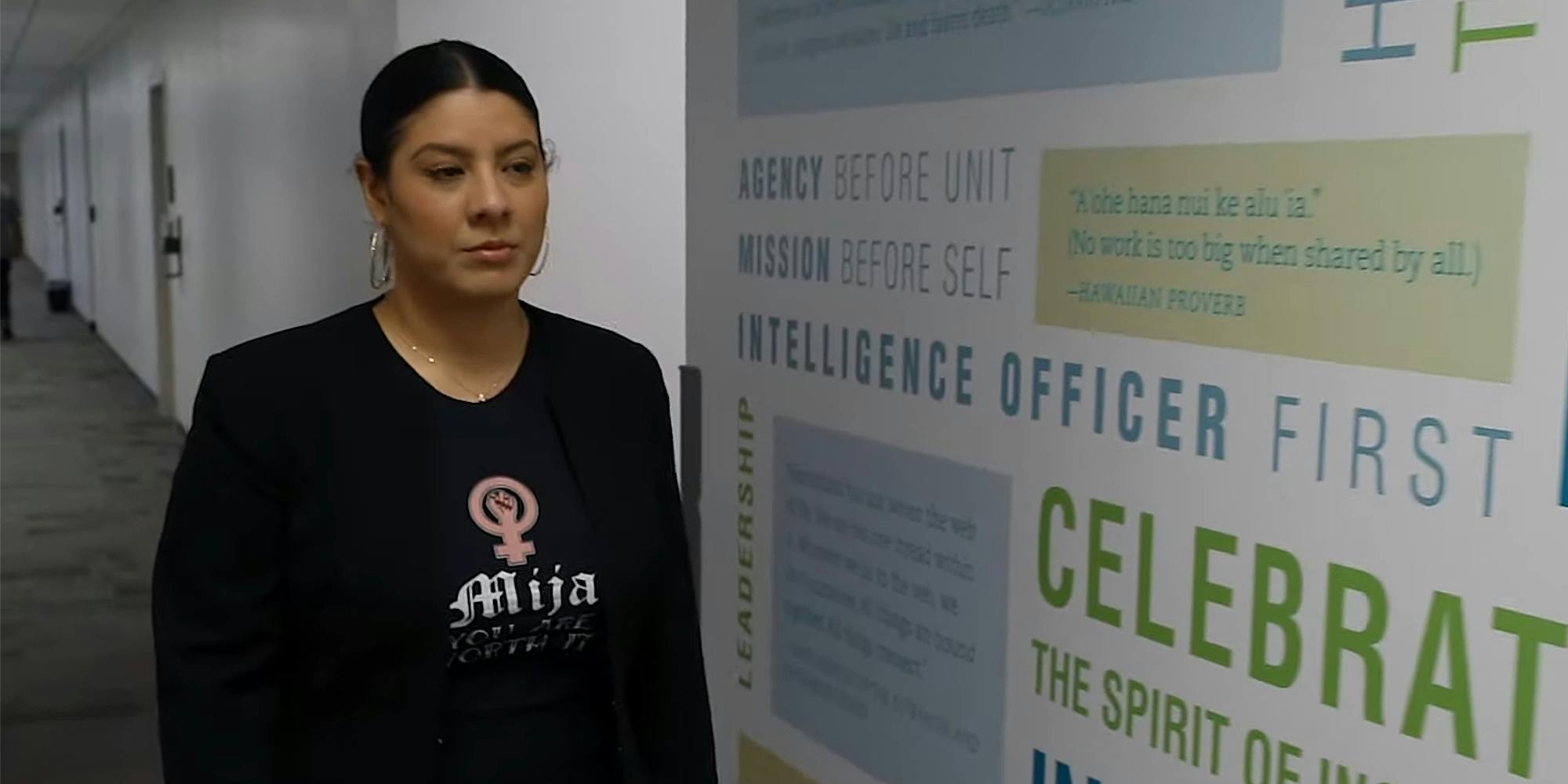 woman in "Mija you are worth it" t-shirt walks past CIA wall art that reads "Agency before unit, Mission before self, Intelligence officer first"