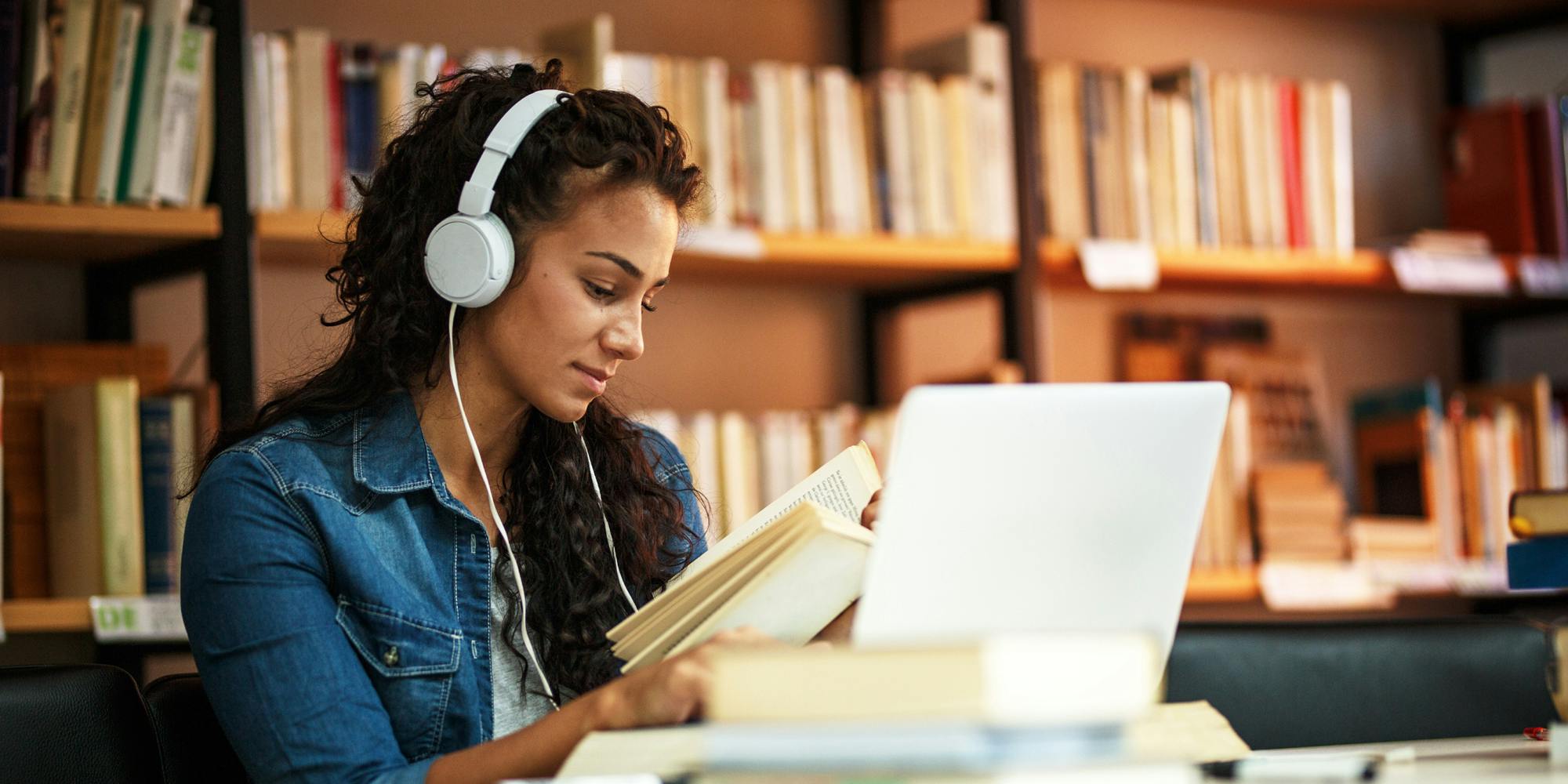 A woman reading a book and working on a laptop inside a library.