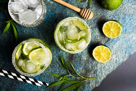 Liquid cannabis in the form of marijuana drinks on a stone table surrounded by weed leaves and limes