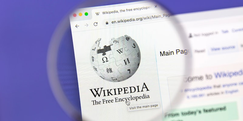 The logo of Wikipedia on a webpage behind a magnifying glass.
