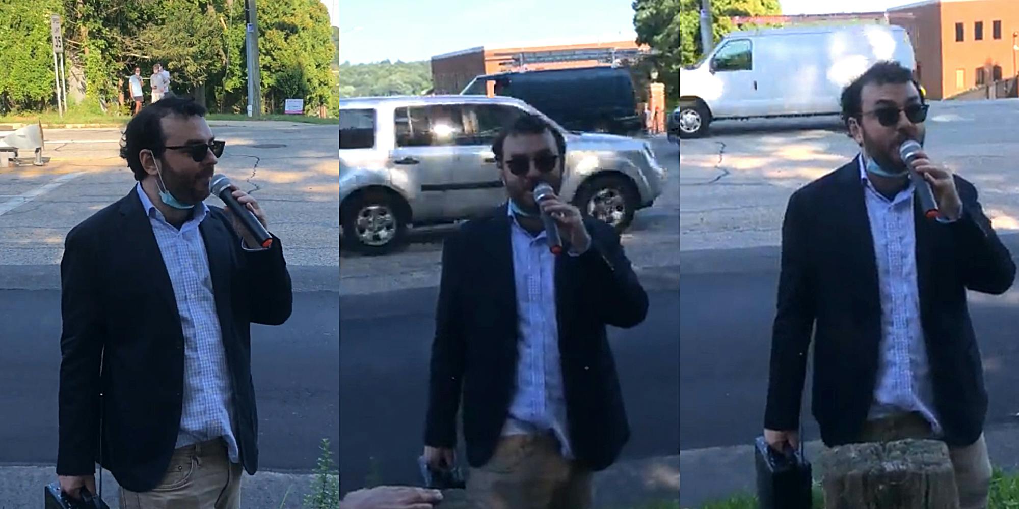 unkempt man in sports jacket with untucked shirt speaks into microphone on sidewalk