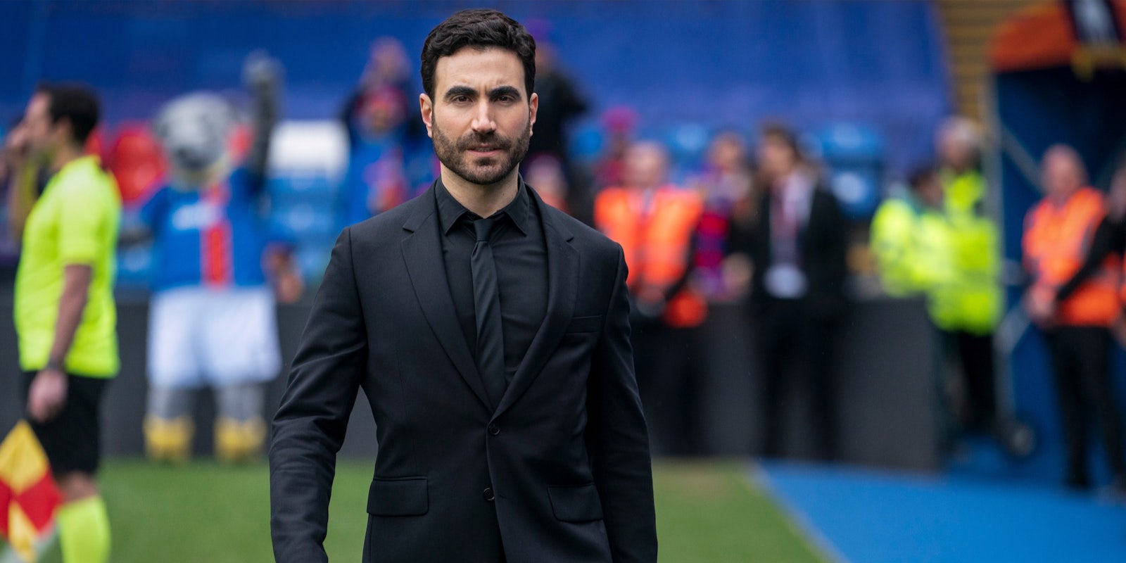 man in black suit walking on to a soccer pitch