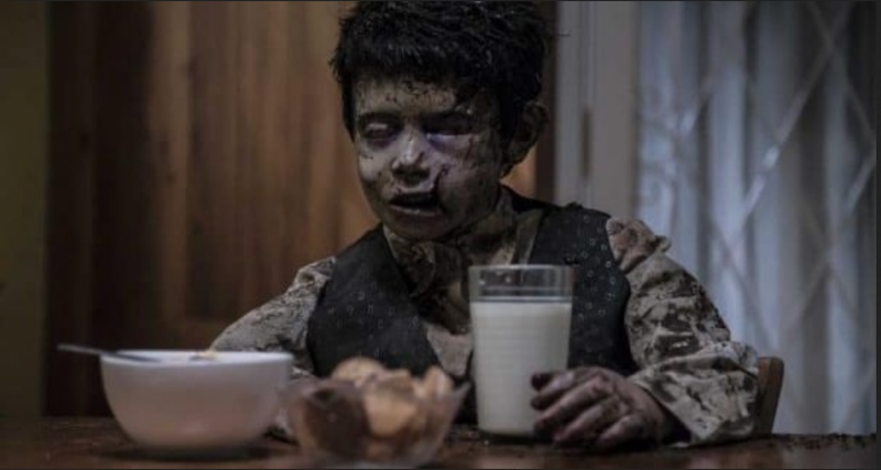 shudder - terrified, a dead boy sits at a table in front of a glass of milk, hiw body clearly rotting after time in the grave