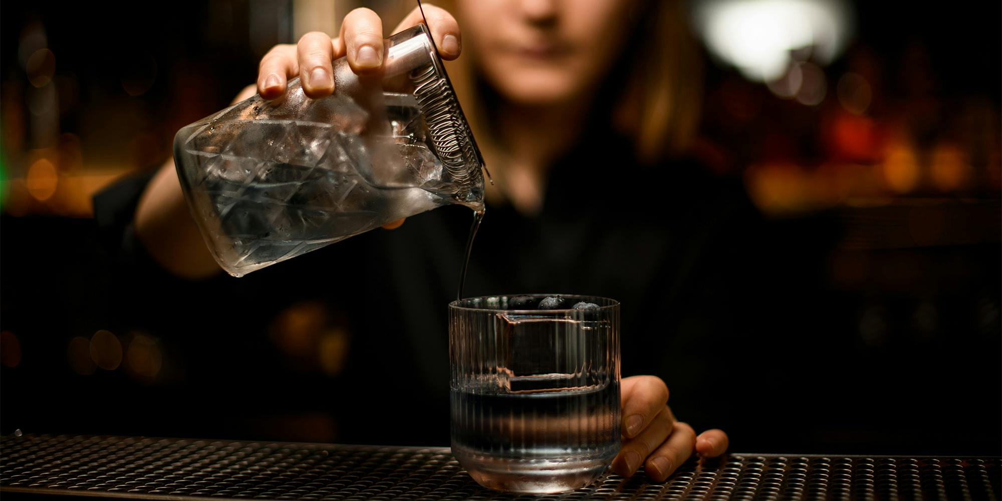 woman bartender holds mixing cup with cocktail and pours it into old-fashioned transparent glass with ice cube and blueberries