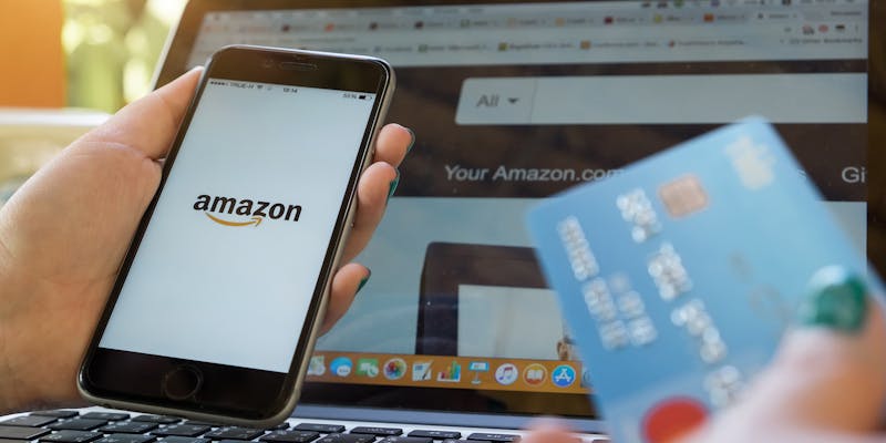 A person holds a phone and credit card while viewing the Amazon website on their computer.