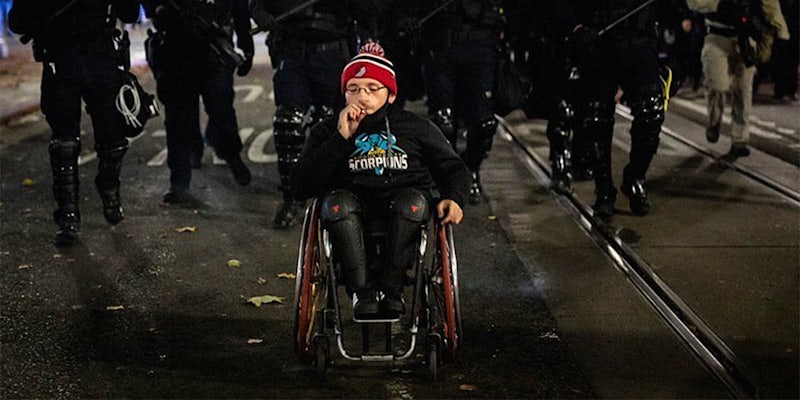 man smoking in wheelchair in front of marching police in riot gear