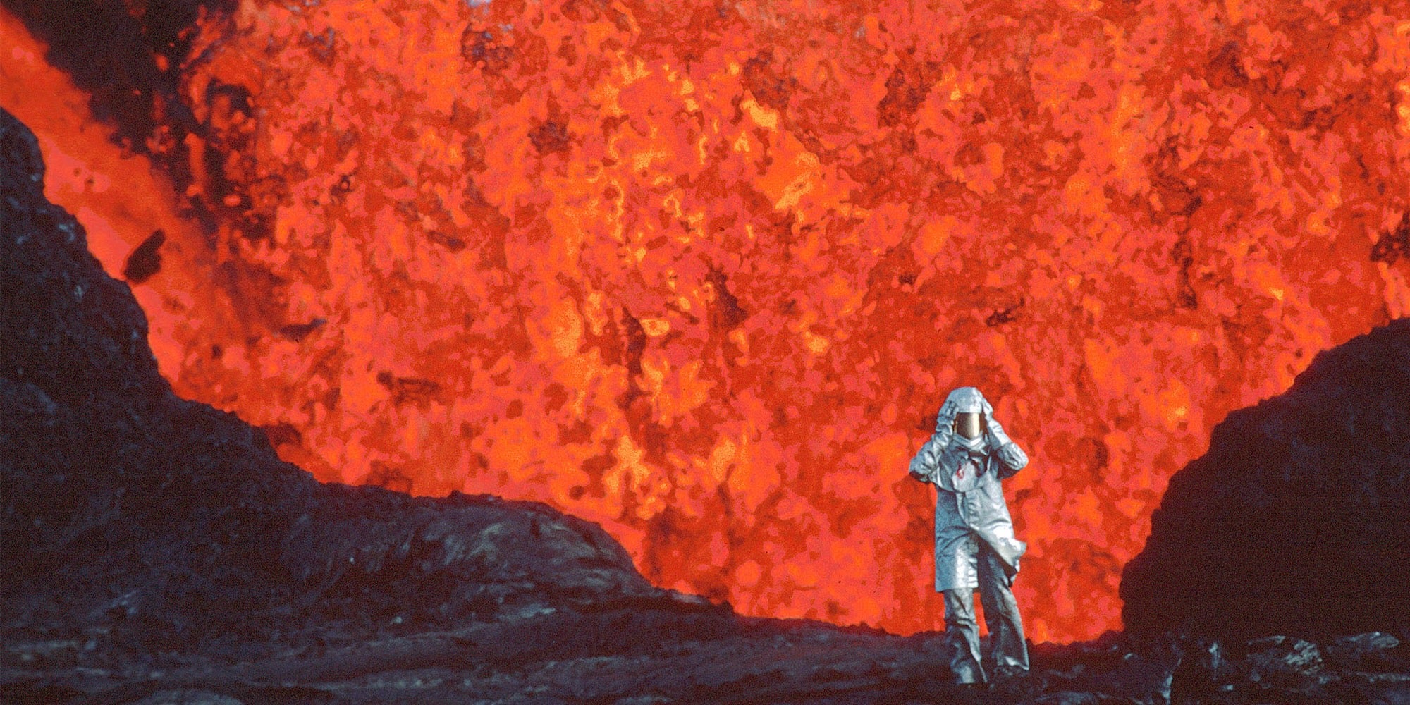 a person in silver gear standing in front of an erupting volcano in fire of love