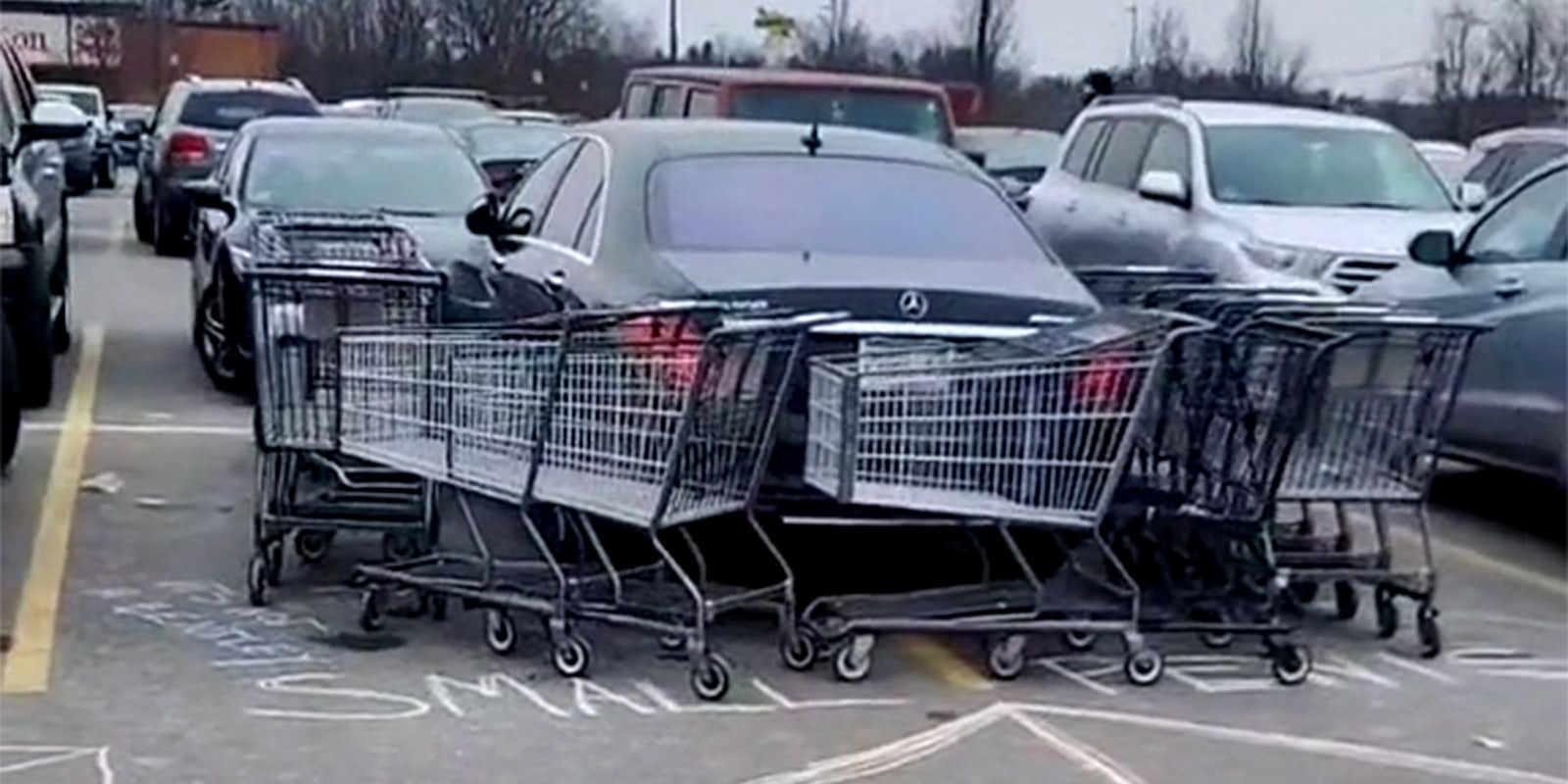 car taking up two spaces surrounded by shopping carts with 'small penis' and an arrow pointing toward the car written in chalk on the ground