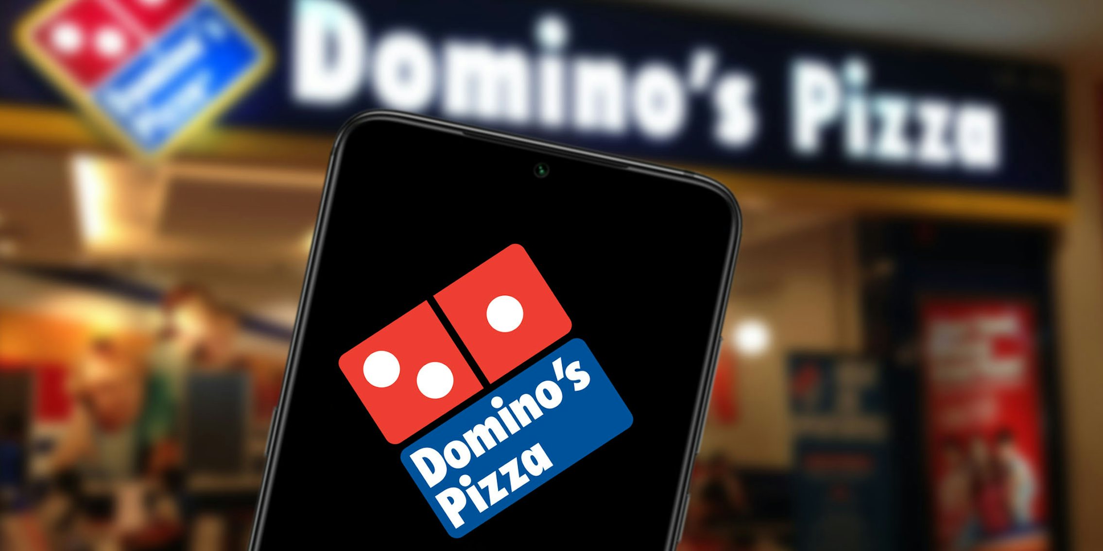 Domino's Pizza shop blurred in background phone in foreground with domino's logo on screen