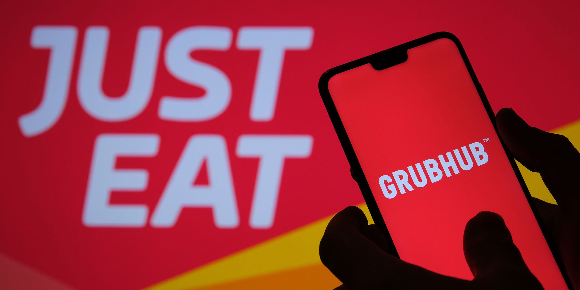 GrubHub app screen on black phone held in hand silhouette with 'Just Eat' blurred in red and yellow background